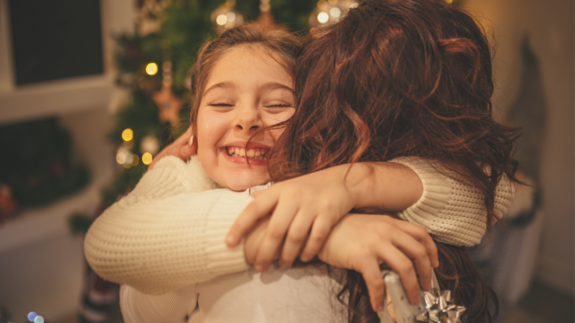 A young girl and her mother sharing a warm hug beside a lit Christmas tree.