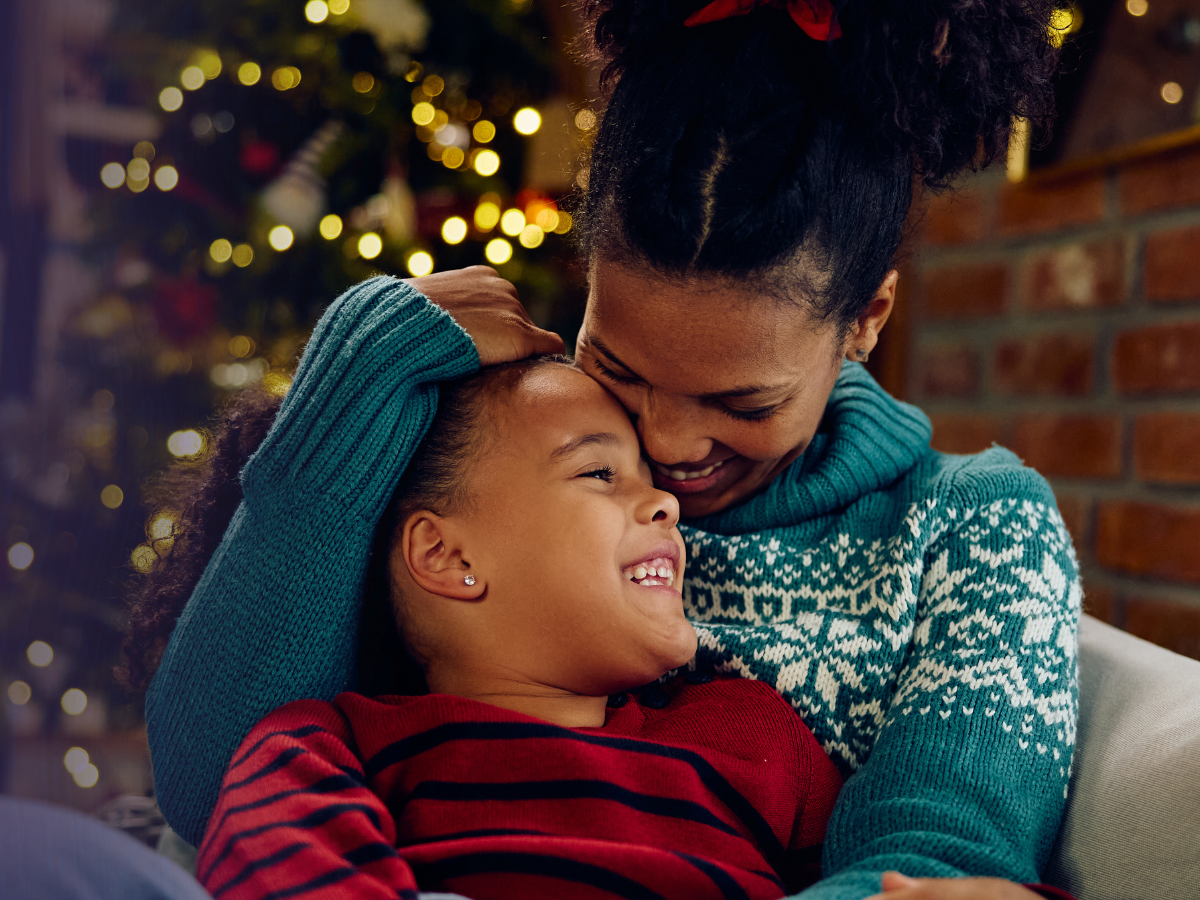 A mother embracing her daughter tenderly by a lit Christmas tree.