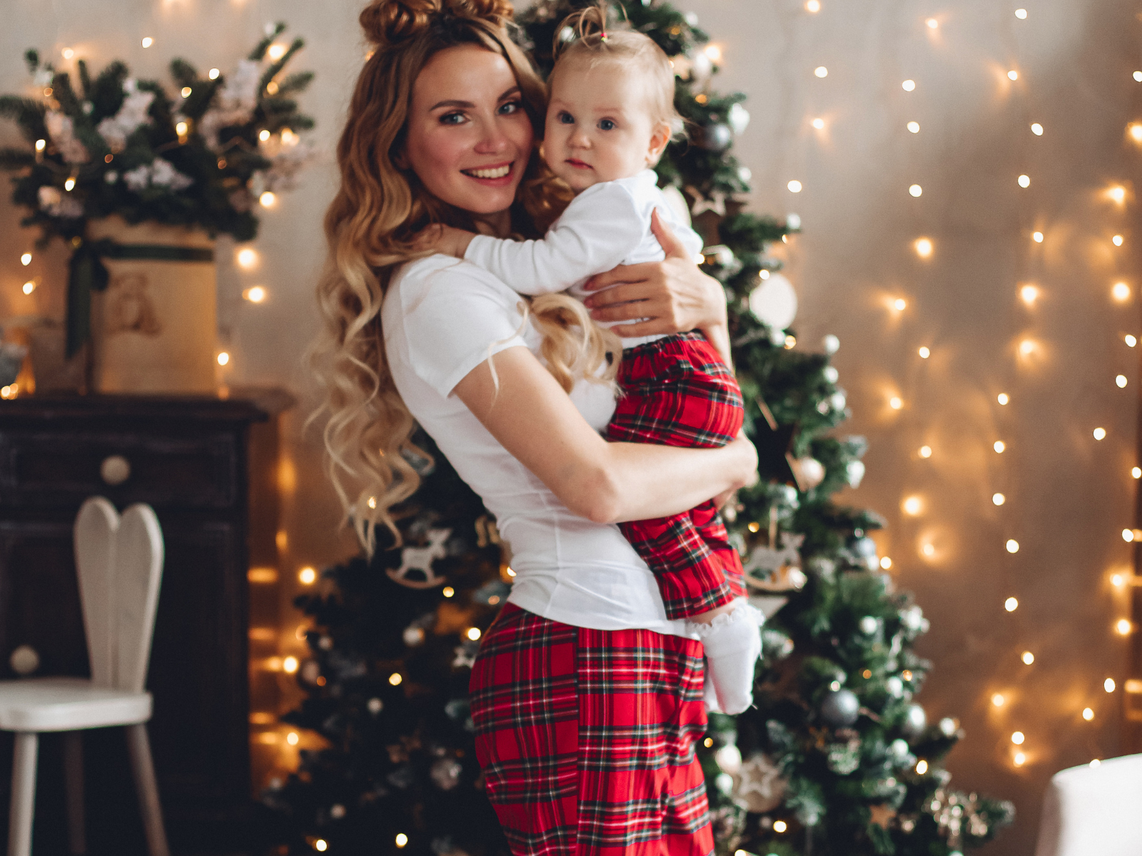 A mother smiling and holding her baby, dressed in holiday pajamas, in front of a Christmas tree.